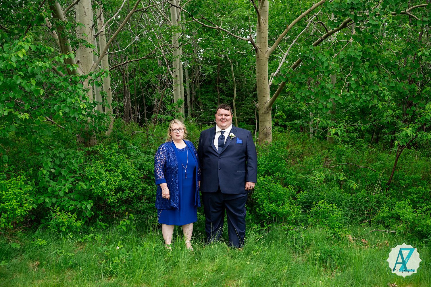 Couple standing together in formal attire on green grass with a wooded forest backdrop.