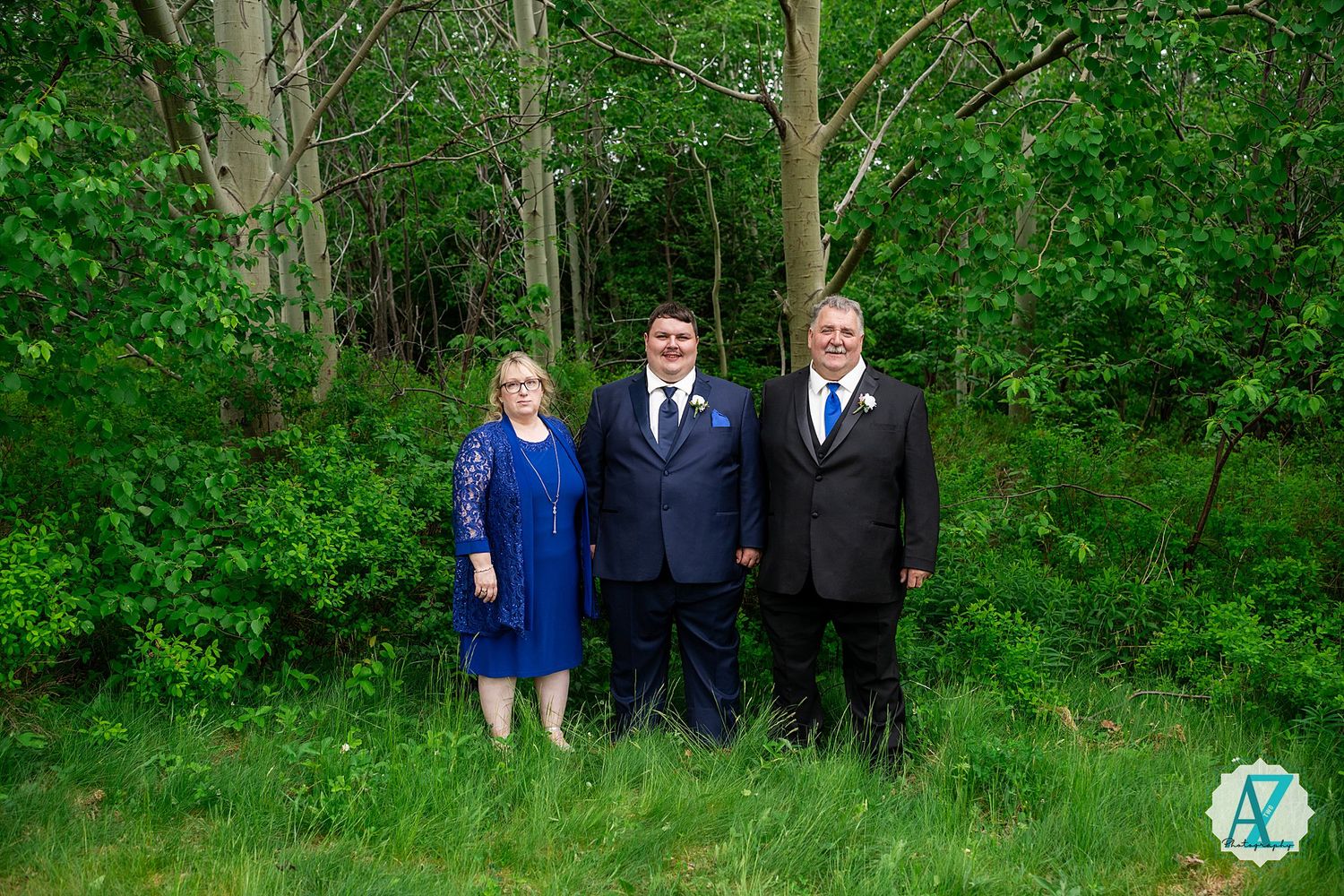 Wedding party members in formal attire pose together against a lush green forest backdrop during an outdoor ceremony.