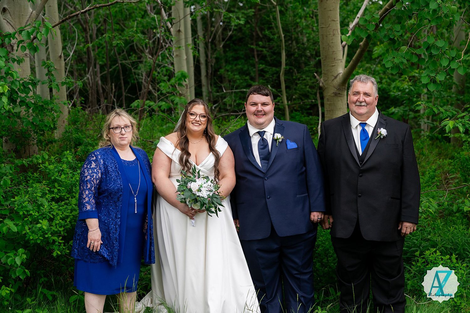 A happy couple in wedding attire poses for a portrait with family members outdoors against a lush green forest backdrop.