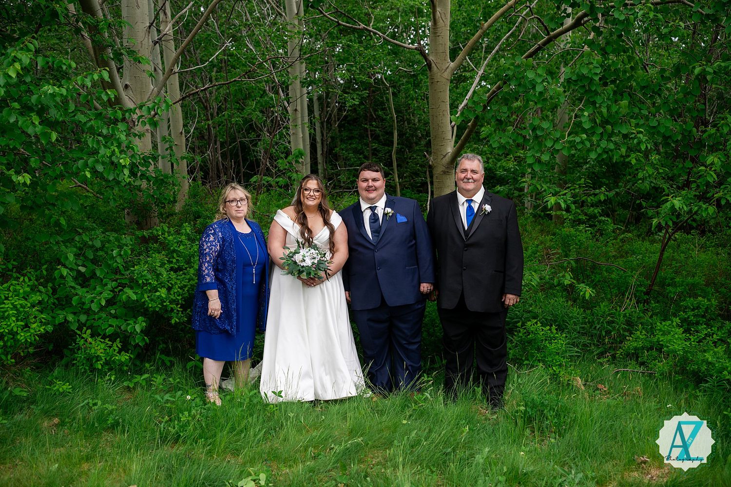 Wedding party in formal attire posing together against a lush green forest backdrop.