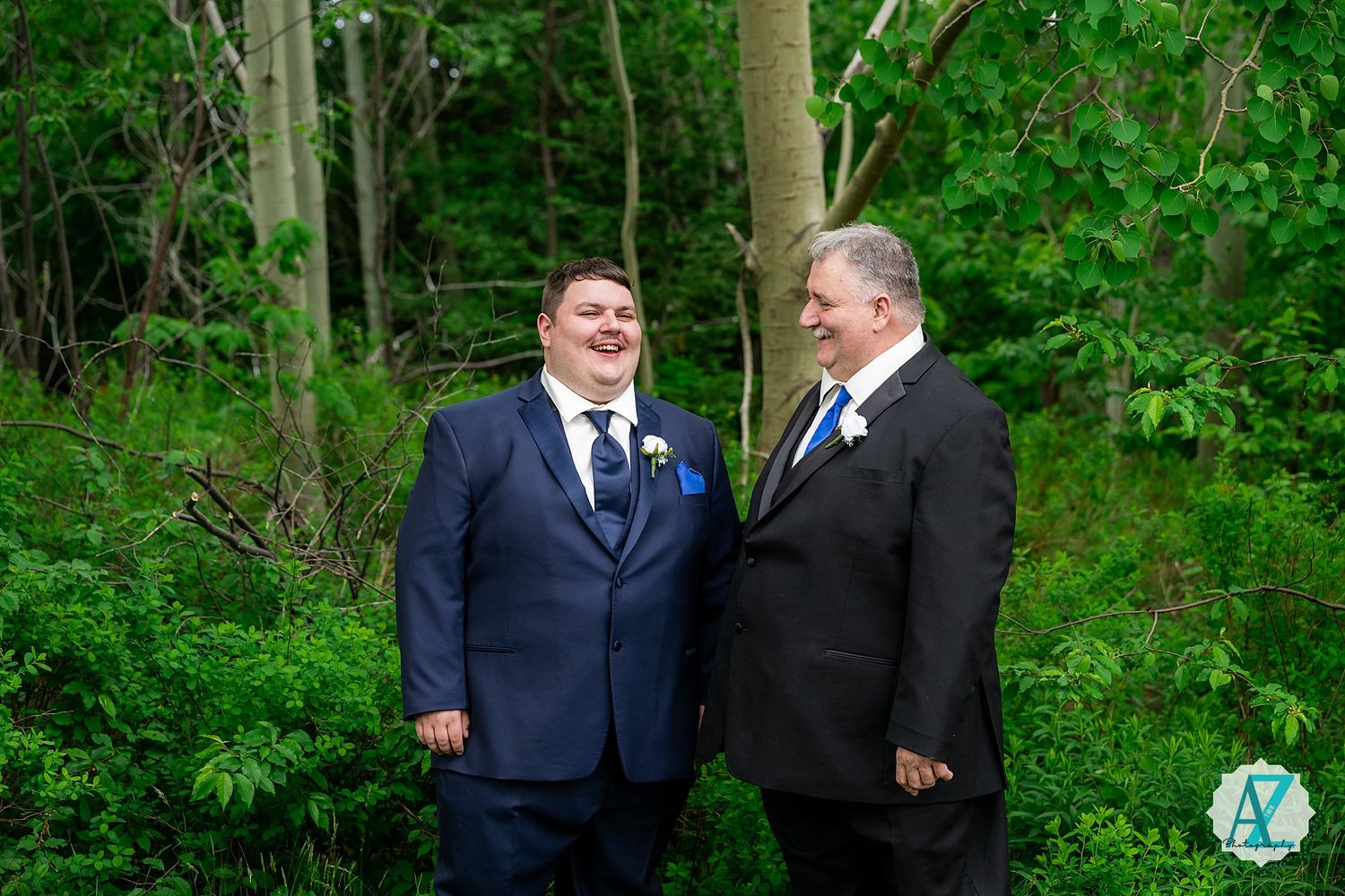 Two people in navy and black suits stand together in a wooded outdoor setting with lush green foliage in the background.