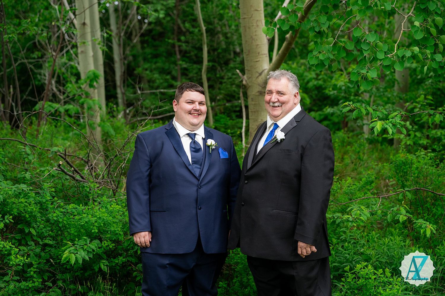 Two men in formal suits stand together for a portrait in front of lush green forest background.