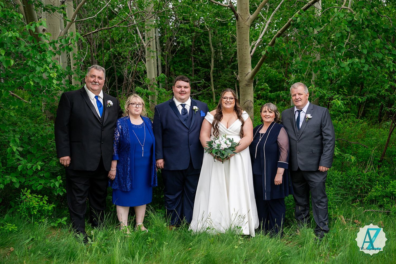 Wedding party dressed in blue and black formal attire poses outdoors in front of green leafy backdrop for photos.