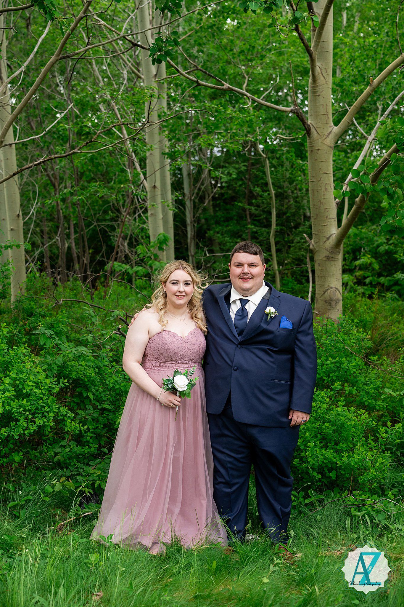 Two people in formal attire stand together in a wooded area with lush green foliage backdrop.