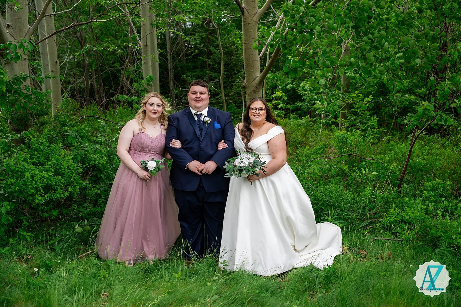Three people pose together outdoors in formal attire with green foliage and trees in the background.