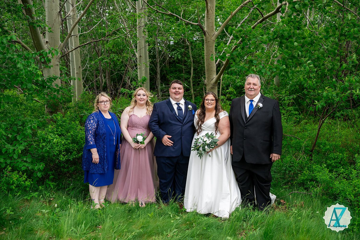 Wedding party wearing formal attire poses in front of lush green forest setting during outdoor celebration.