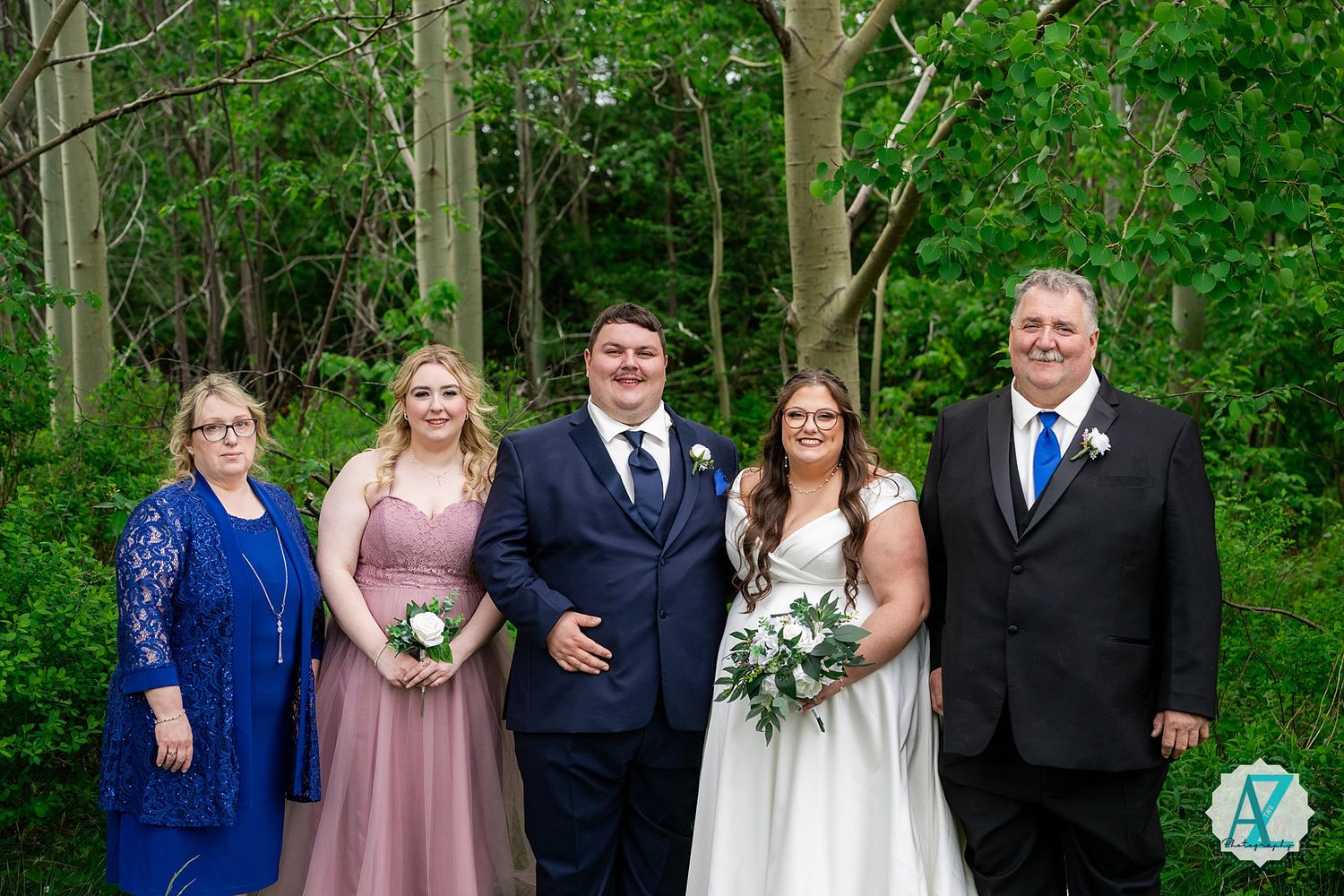 Wedding party in formal attire poses together against lush green forest background during outdoor ceremony.