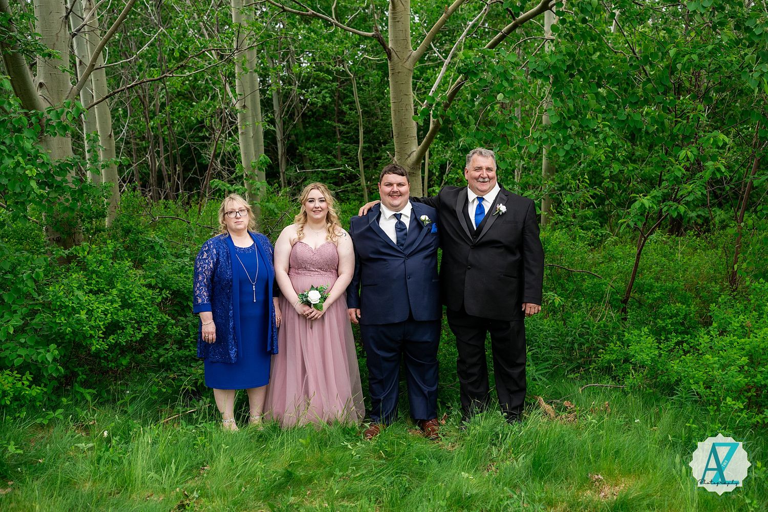 A group in formal attire poses together against a lush green forest backdrop during an outdoor event.
