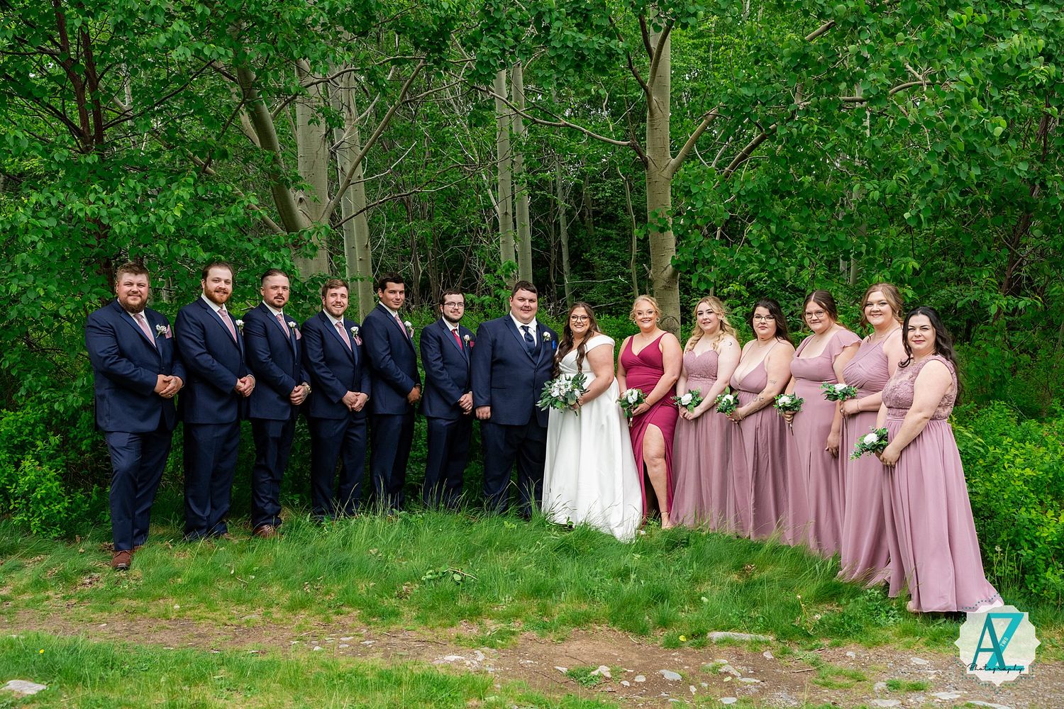 Wedding party in formal attire poses outdoors with blue suits and pink dresses against a lush green forest backdrop.