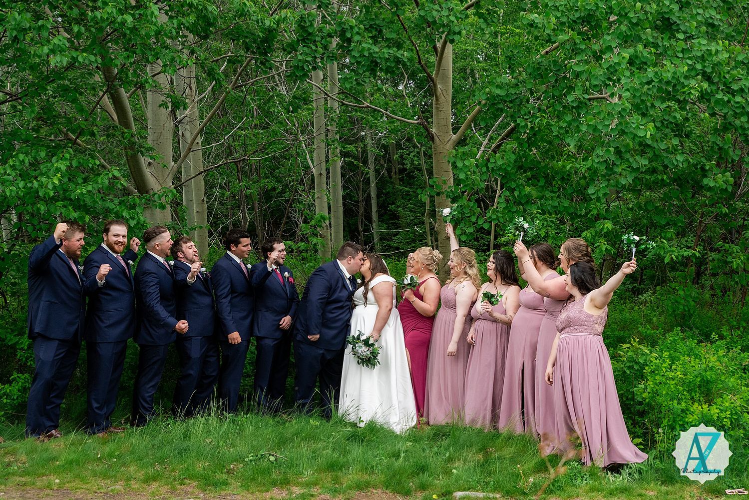 Wedding party in formal attire poses outdoors against lush green forest backdrop in cascading line formation.