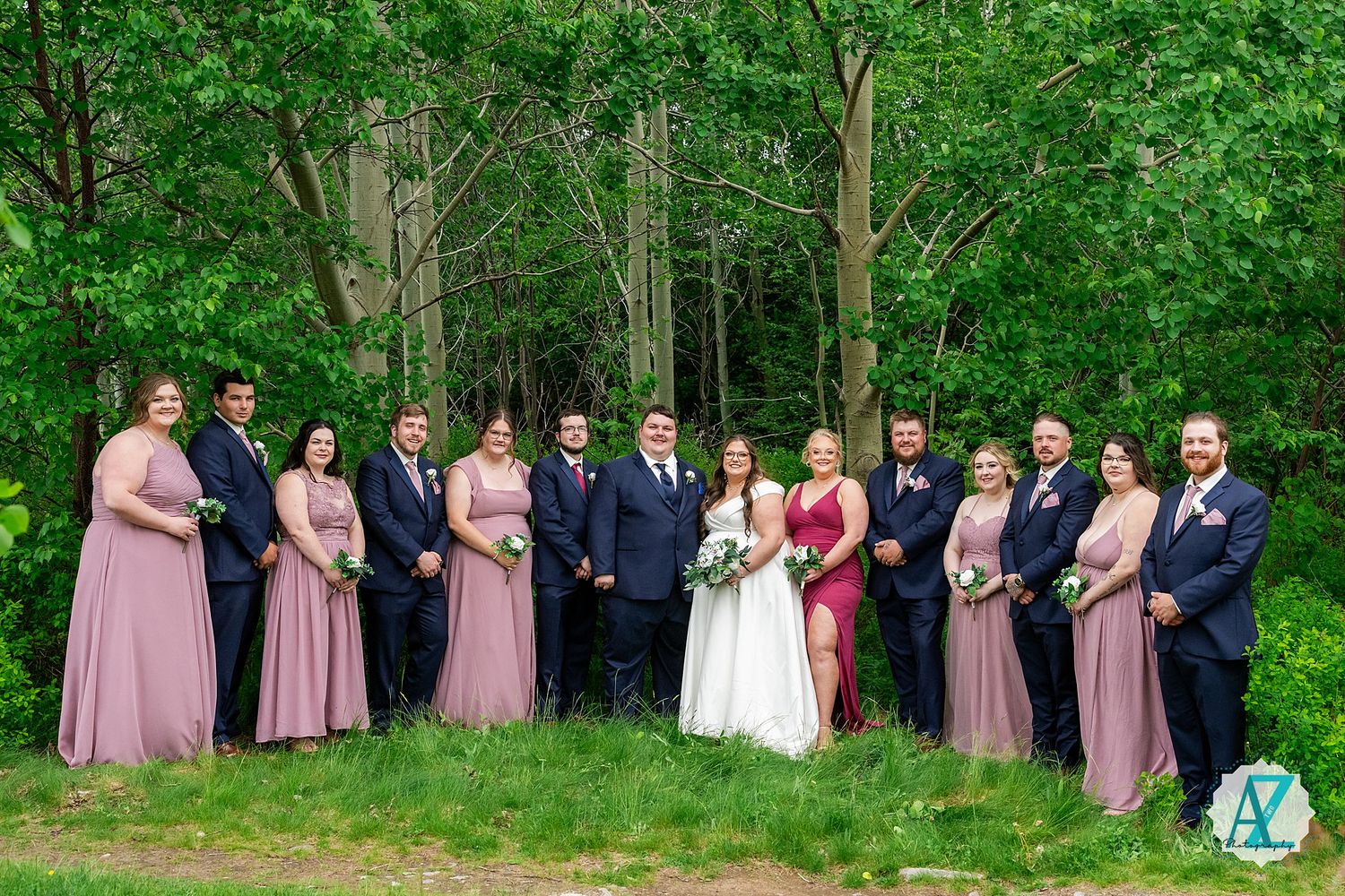Wedding party poses together outdoors in pink and navy attire against lush green forest backdrop.
