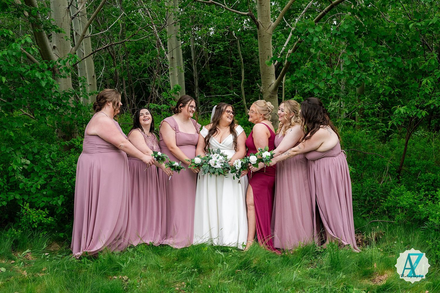 Wedding bridesmaids wearing pink dresses pose playfully on grass in front of lush green forest backdrop.
