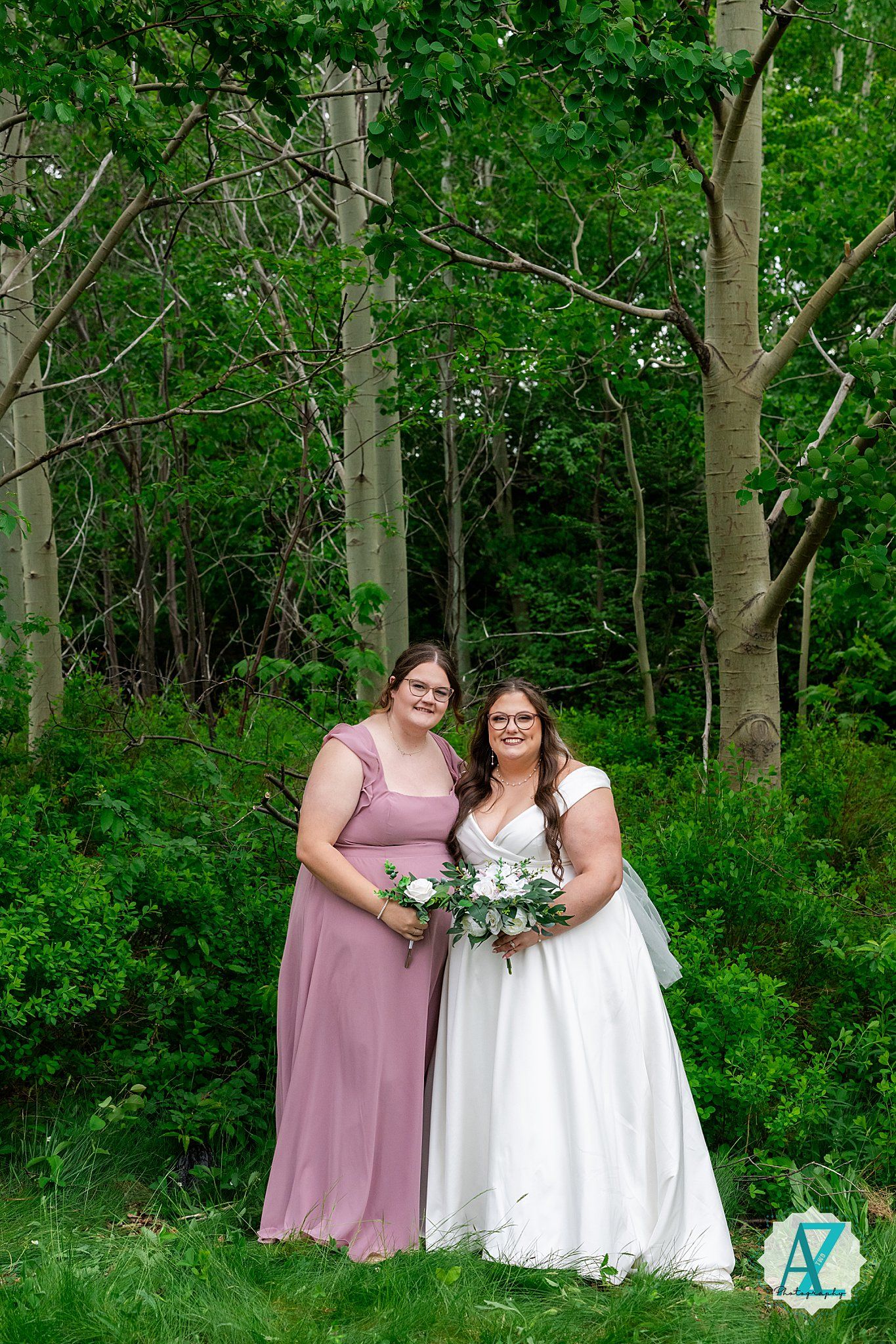 Wedding photo taken in a lush green forest setting with two people in mauve and white gowns holding floral bouquets.