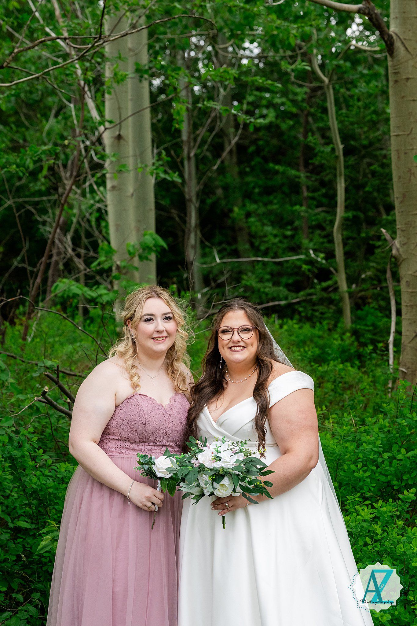 A bridal party in dusty rose and white dresses poses together in a lush green forest setting.