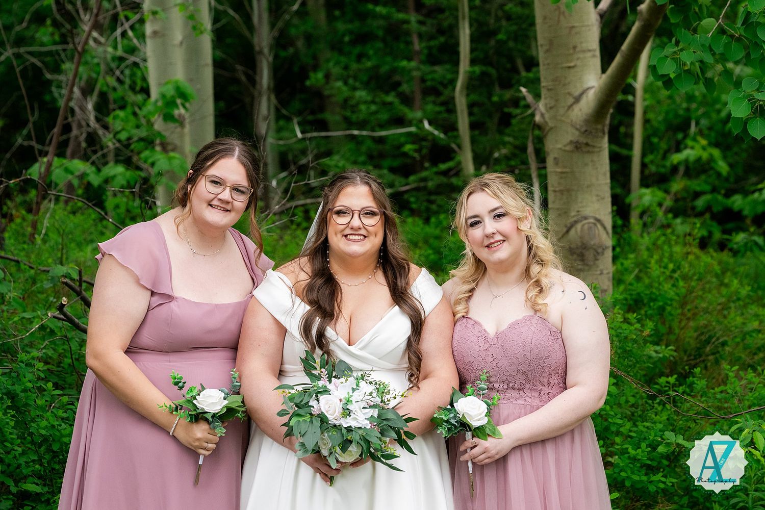 Three women in pink and floral dresses pose together outdoors in front of lush green trees while holding bouquets.