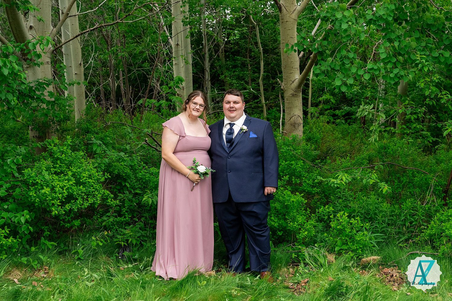 Couple in formal attire takes romantic outdoor photos in a lush green forest setting.
