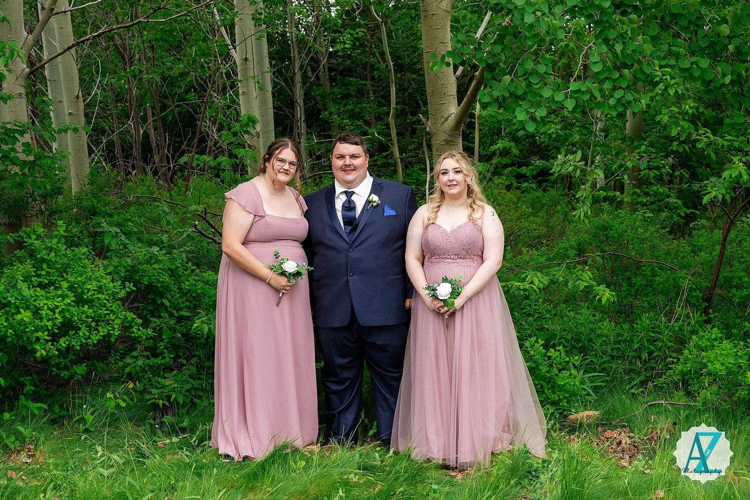 A wedding party outdoors with two members in pink dresses and one in a navy suit standing in front of greenery.