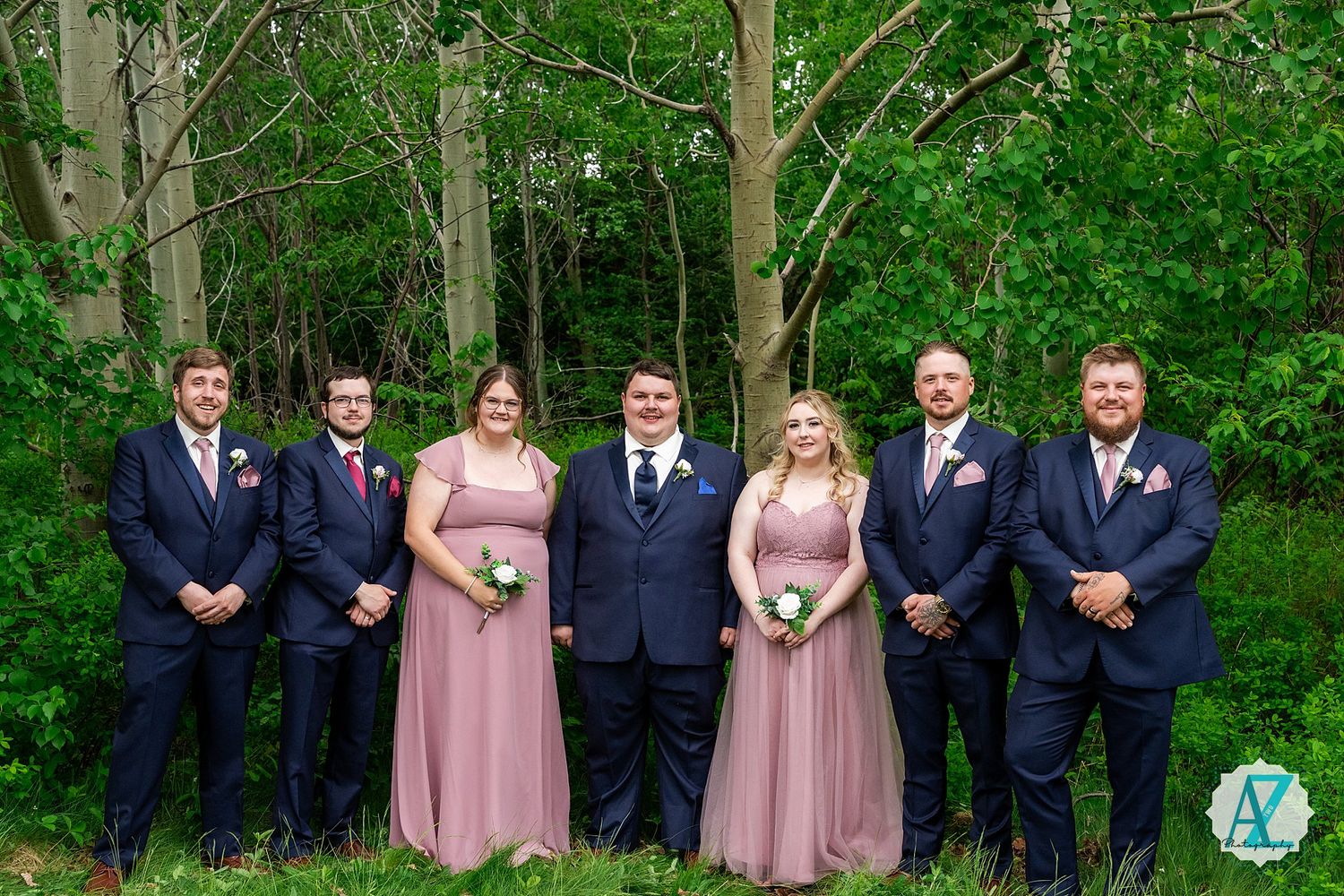 Wedding party in navy suits and dusty rose gowns stand together against a wooded green backdrop.