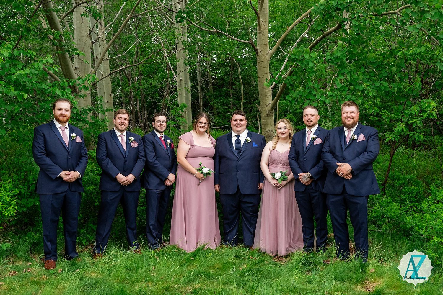 A formal wedding group photo showing bridesmaids in pink dresses and groomsmen in dark suits against a wooded backdrop.