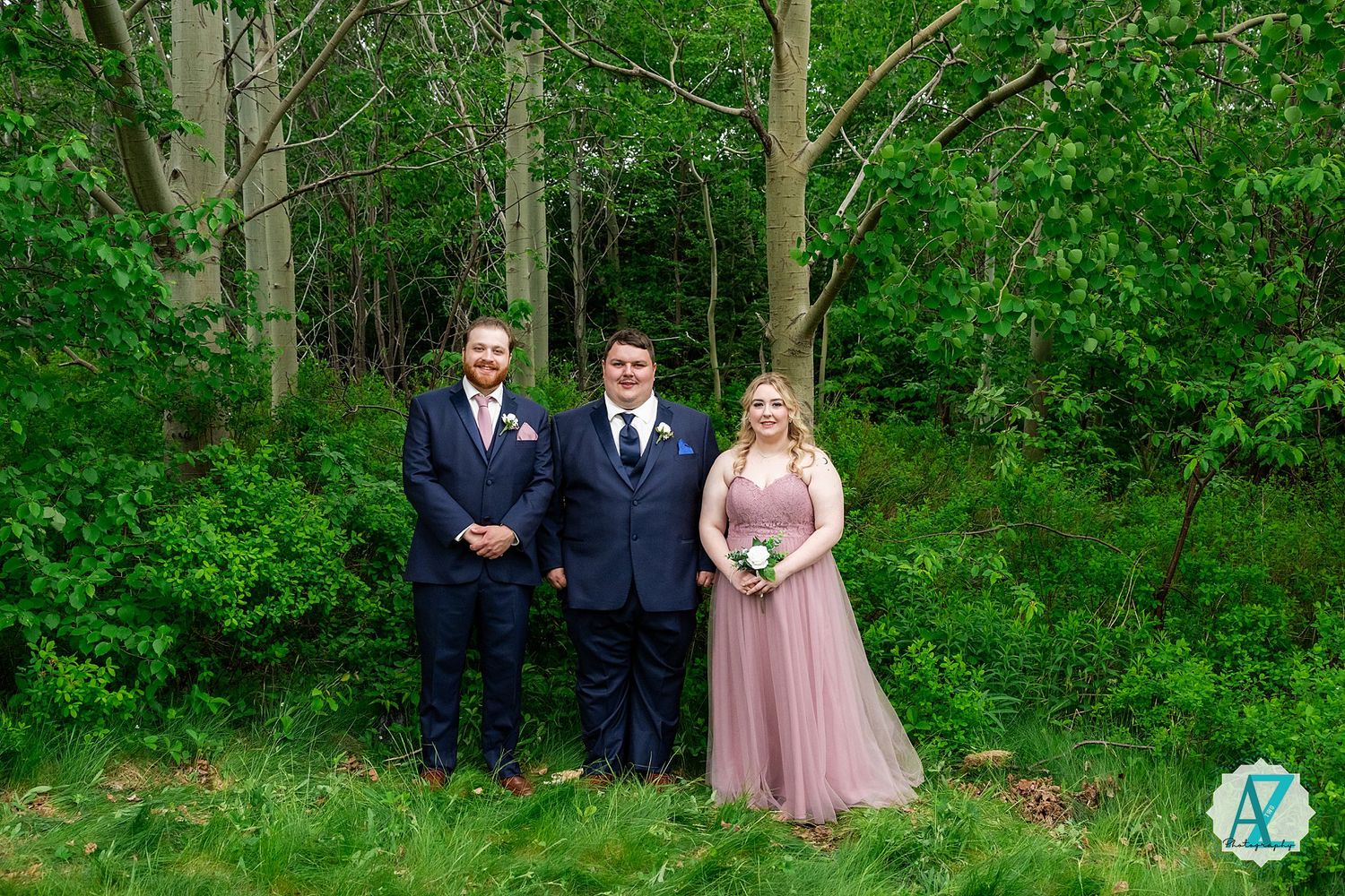 Three wedding attendants in formal attire pose together against a lush green forest backdrop.