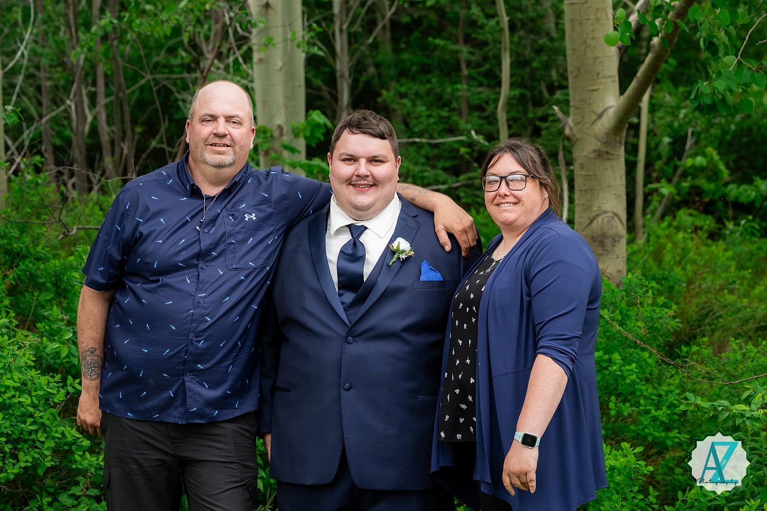 Three people standing together outdoors in formal attire against a green forest background.