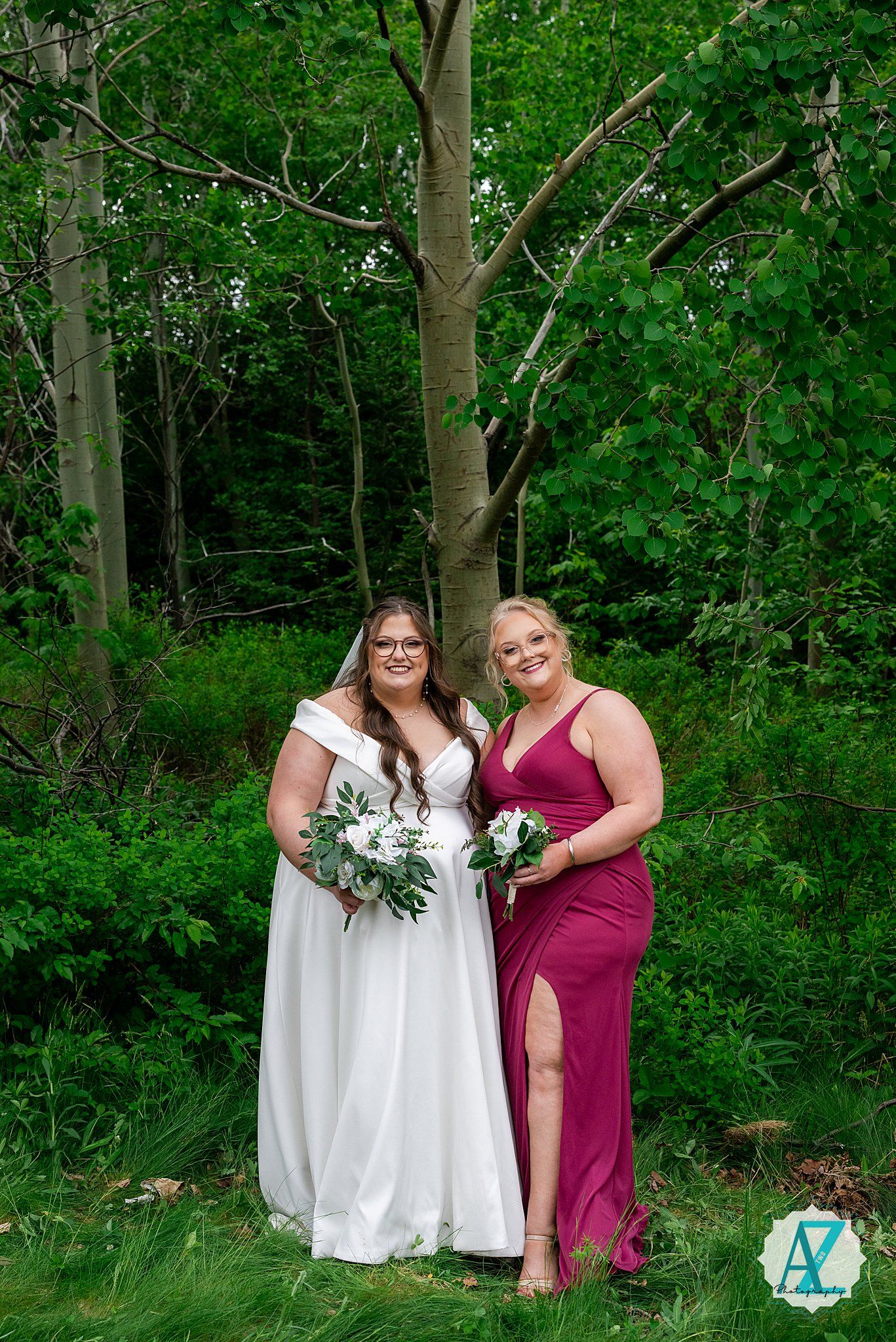 Two women pose in formal dresses with bouquets against a woodland backdrop, one in white and one in burgundy.