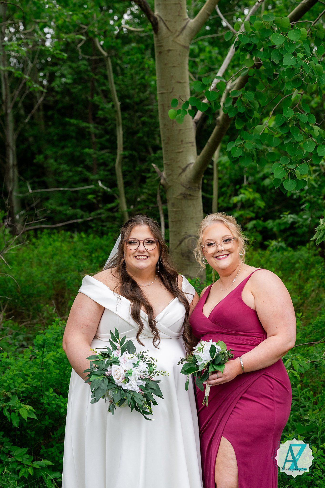 Two people in formal attire pose together outdoors, one in a white wedding dress and one in a burgundy gown holding bouquets.