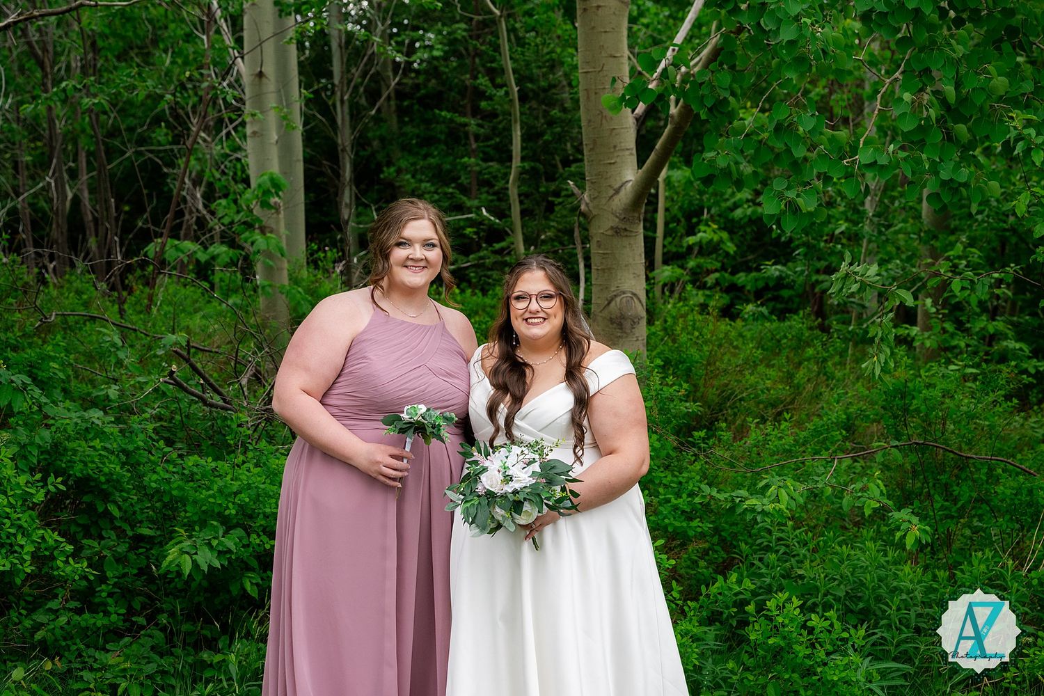 Two people wearing pink bridesmaid dress and white wedding gown pose together in front of green forest backdrop.