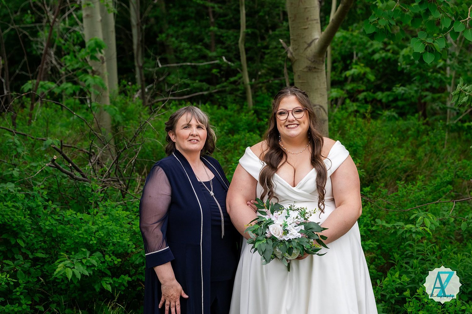 Two people stand together in a woodland setting with lush green foliage, one wearing navy and one in a white dress with flowers.