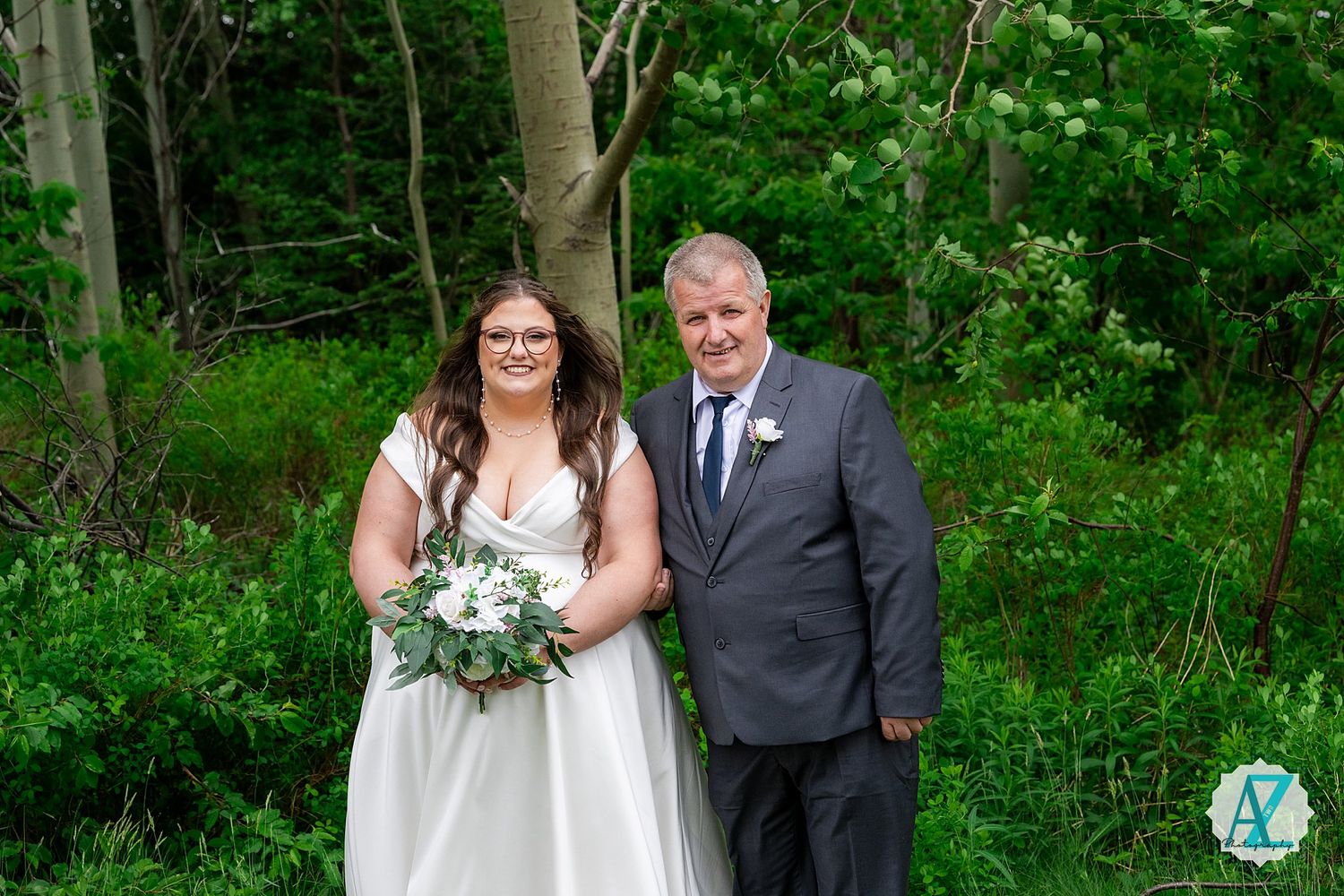 Two people in wedding attire stand together in a lush green wooded setting holding a bouquet of flowers.