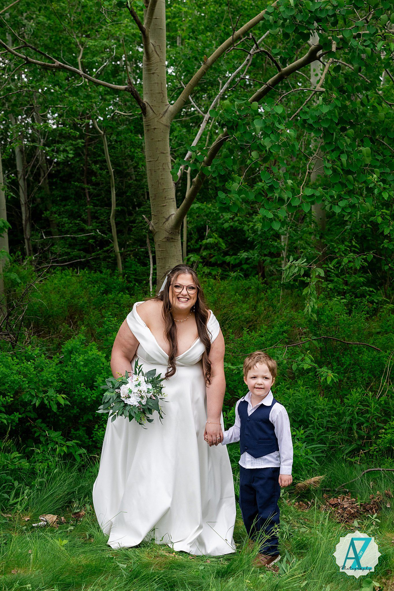 A person in a white wedding dress stands next to a young child in formal attire among green trees and foliage.