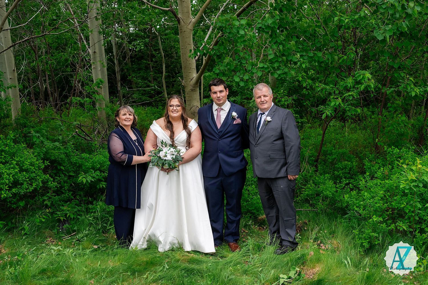 Wedding party members pose together in front of lush green forest scenery during outdoor celebration.