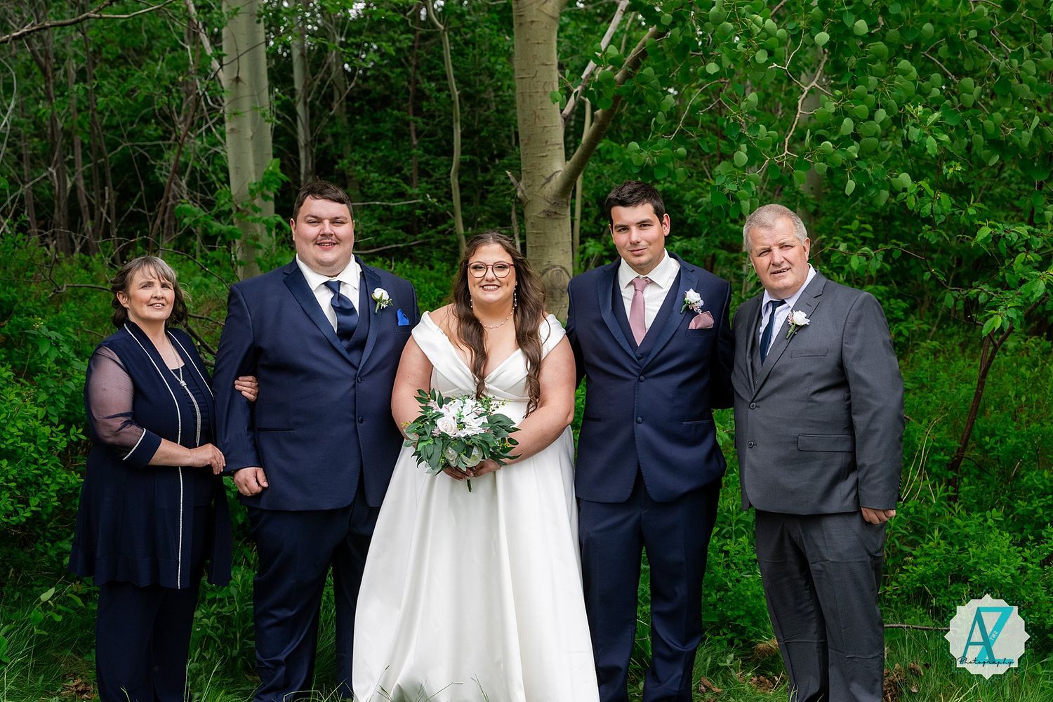 A wedding party poses together in front of lush green foliage at an outdoor ceremony.