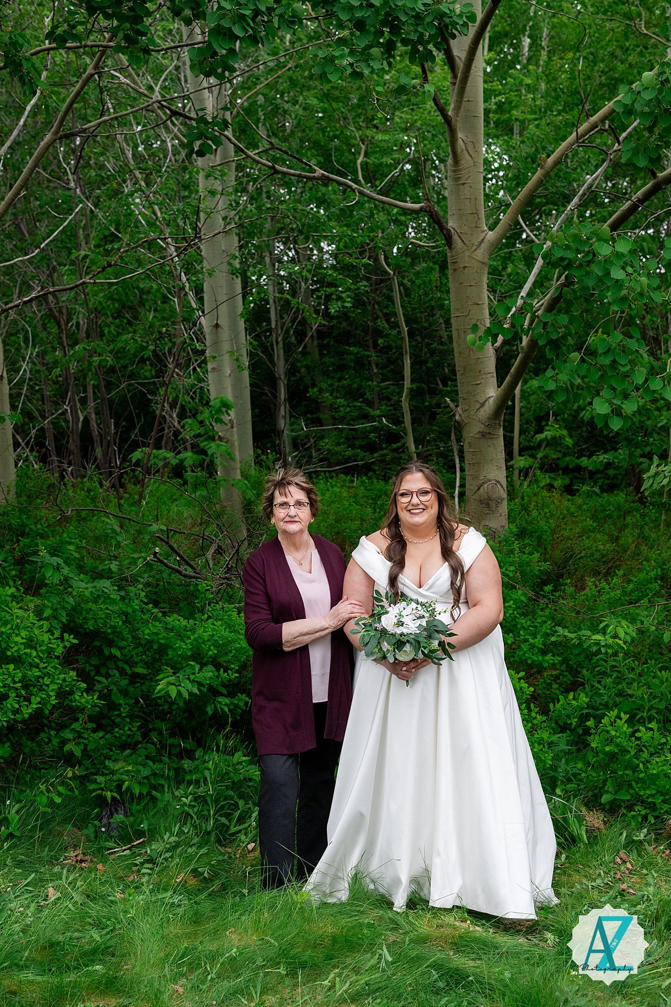 A wedding photo in a lush green forest setting with a couple posing together, one wearing white and holding a bouquet.