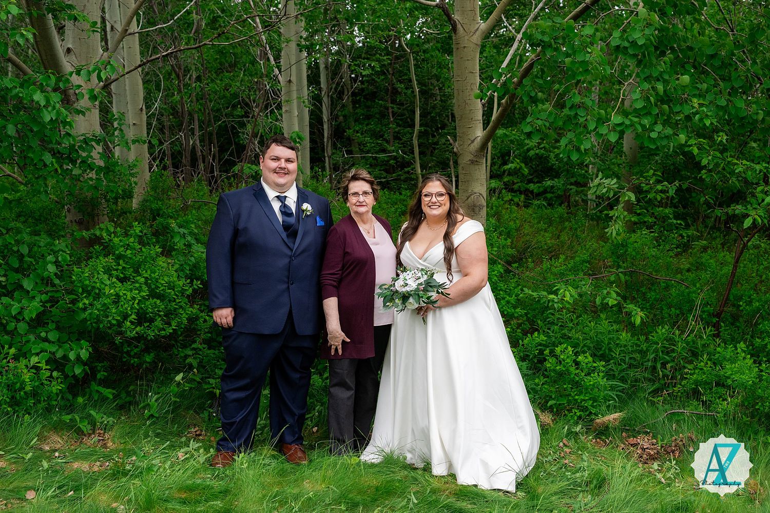 Three people pose together against a green forest backdrop at an outdoor wedding ceremony.