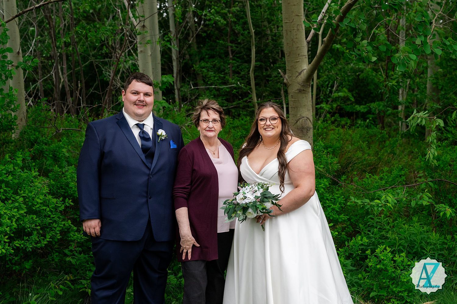 Three people pose together in a wooded outdoor setting with greenery and trees in the background.