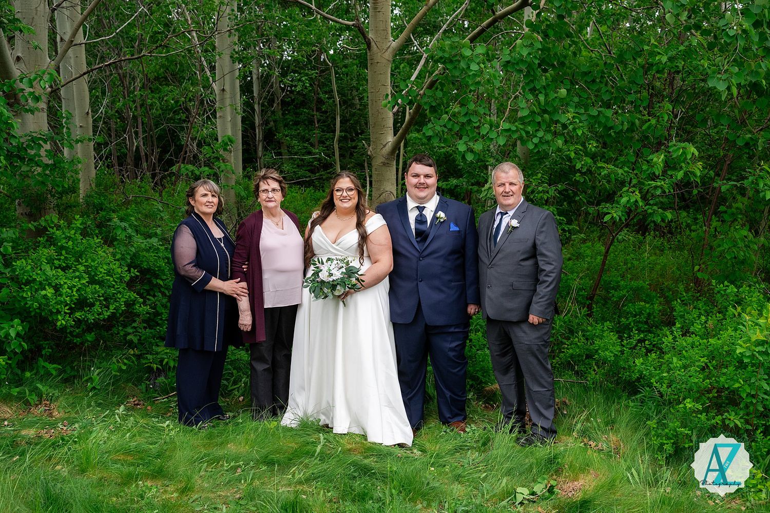 A wedding party poses outdoors in front of lush green forest vegetation during a springtime celebration.