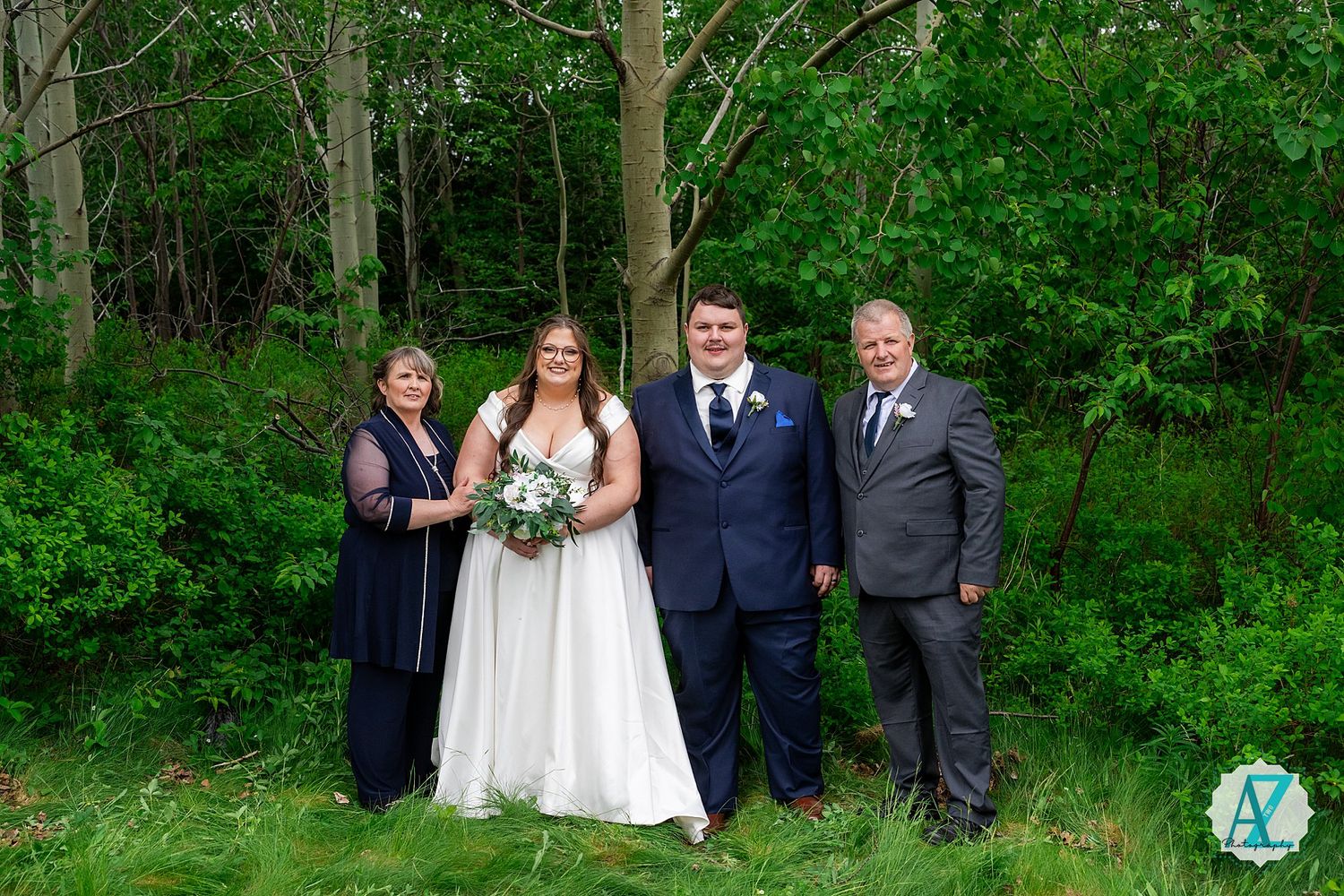 Wedding party poses in front of lush green forest backdrop on summer day.