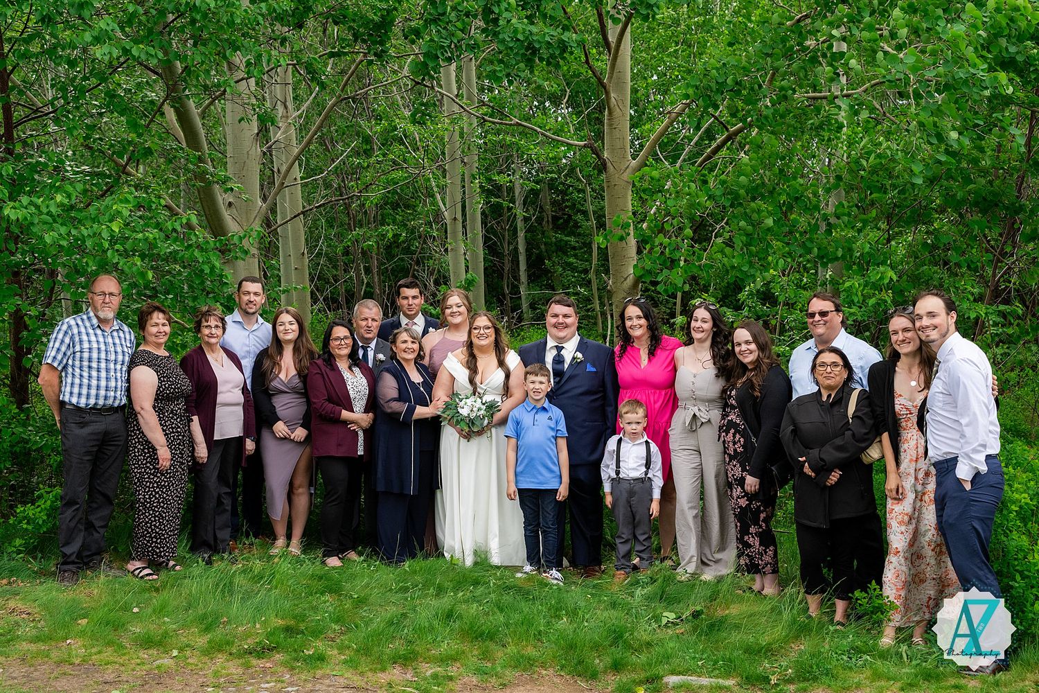 A group of wedding guests standing in a line outdoors with lush green trees and foliage in the background.