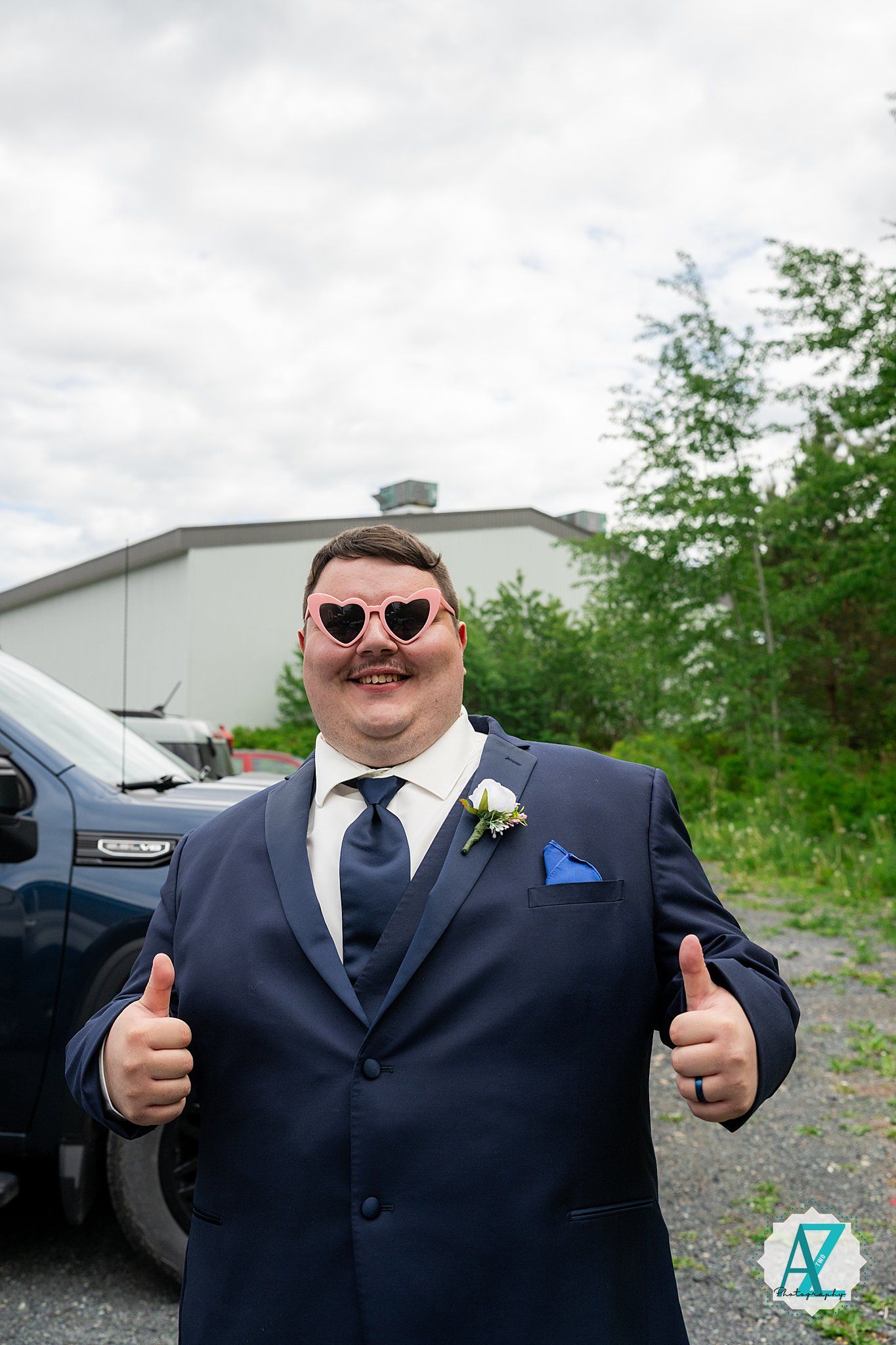 Someone wearing sunglasses and a navy suit with a boutonniere gives two thumbs up while smiling on their wedding day.