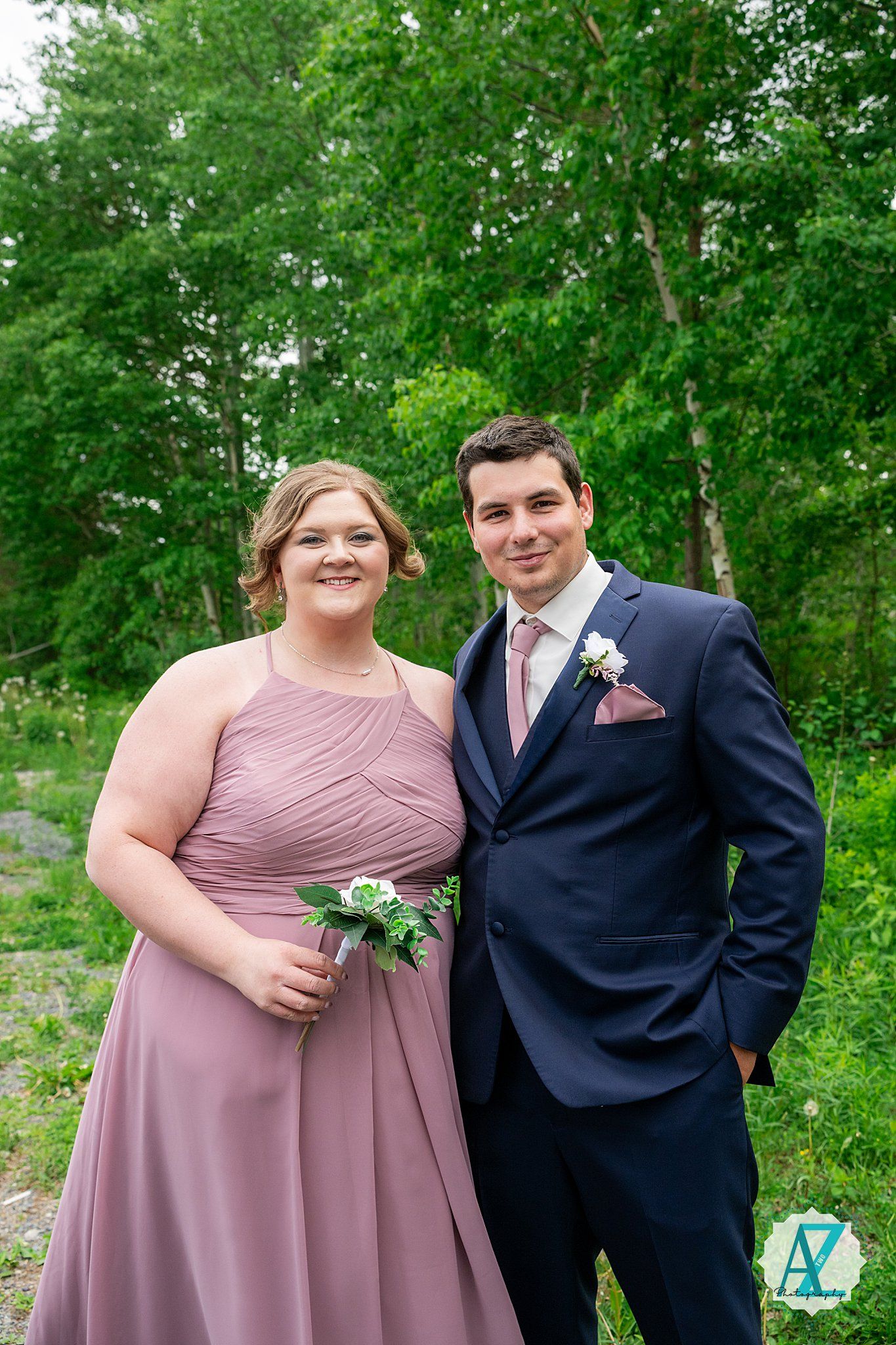 A couple dressed in formal wedding attire with a dusty rose gown and navy suit pose together outdoors among green trees.