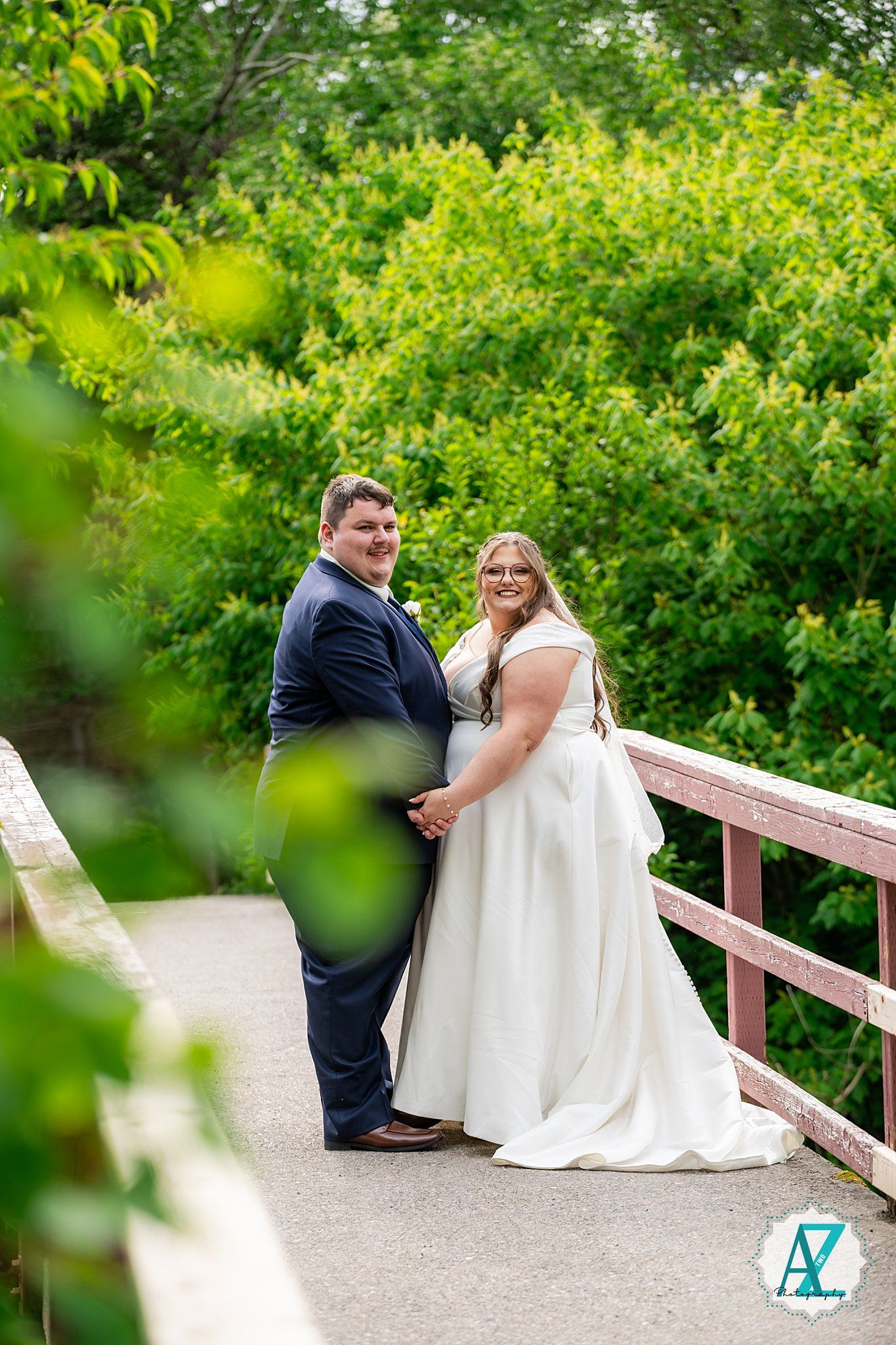 A couple poses on a wooden bridge surrounded by lush green foliage during a sunny outdoor wedding photoshoot.