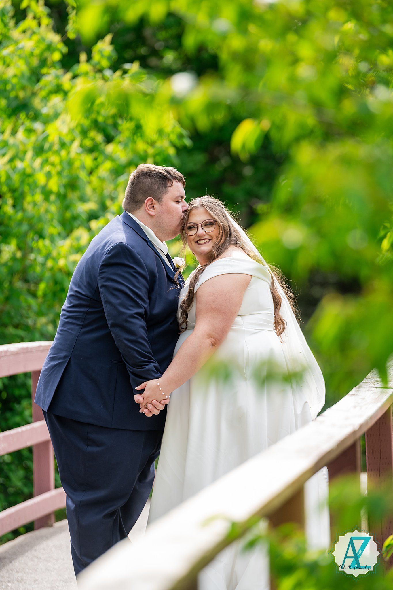 A couple shares a romantic moment on a wooden bridge surrounded by lush green foliage on their wedding day.