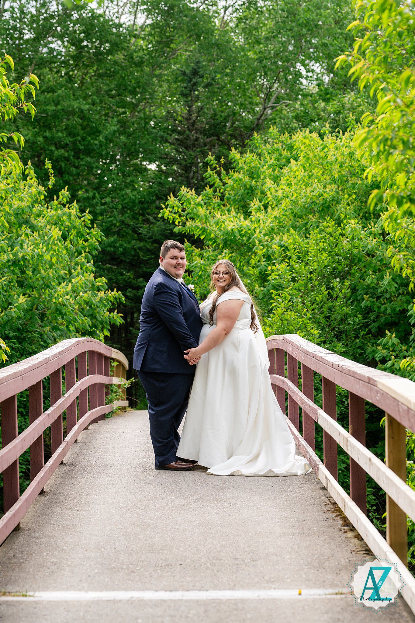 Two people in formal wedding attire stand together on a wooden bridge surrounded by lush green foliage.