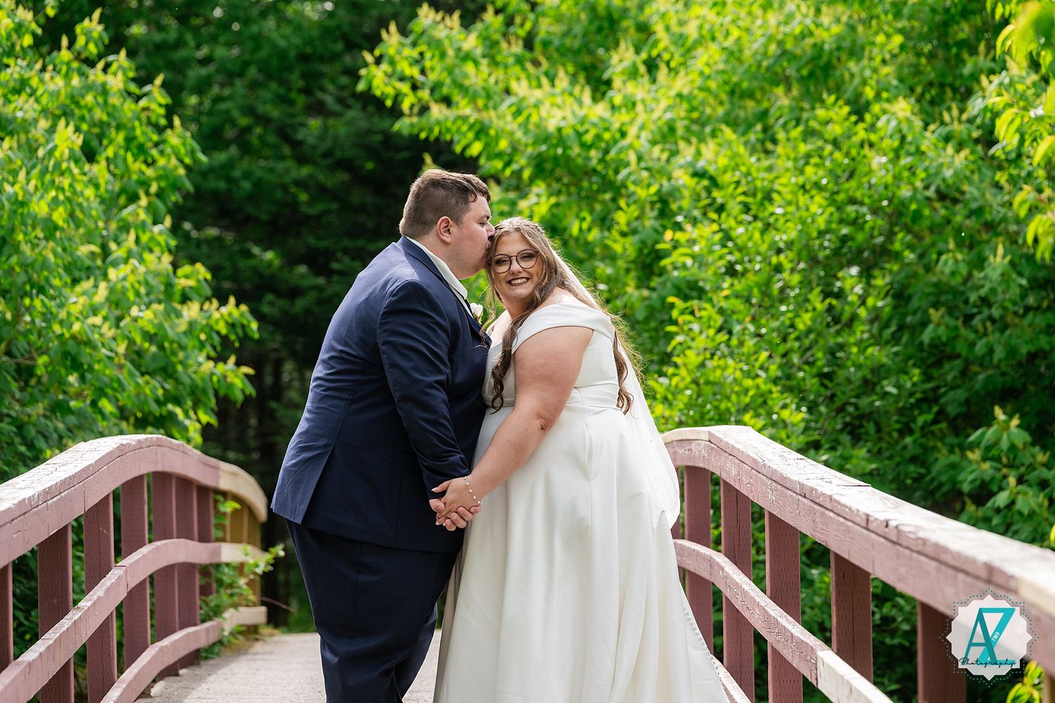 Newlywed couple standing on wooden bridge surrounded by lush green foliage during outdoor summer wedding.