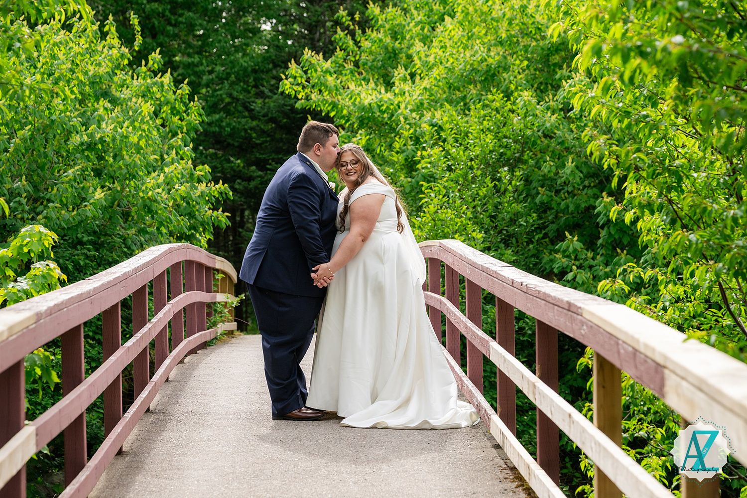 A couple in wedding attire share a romantic moment while standing on a wooden bridge surrounded by lush greenery.