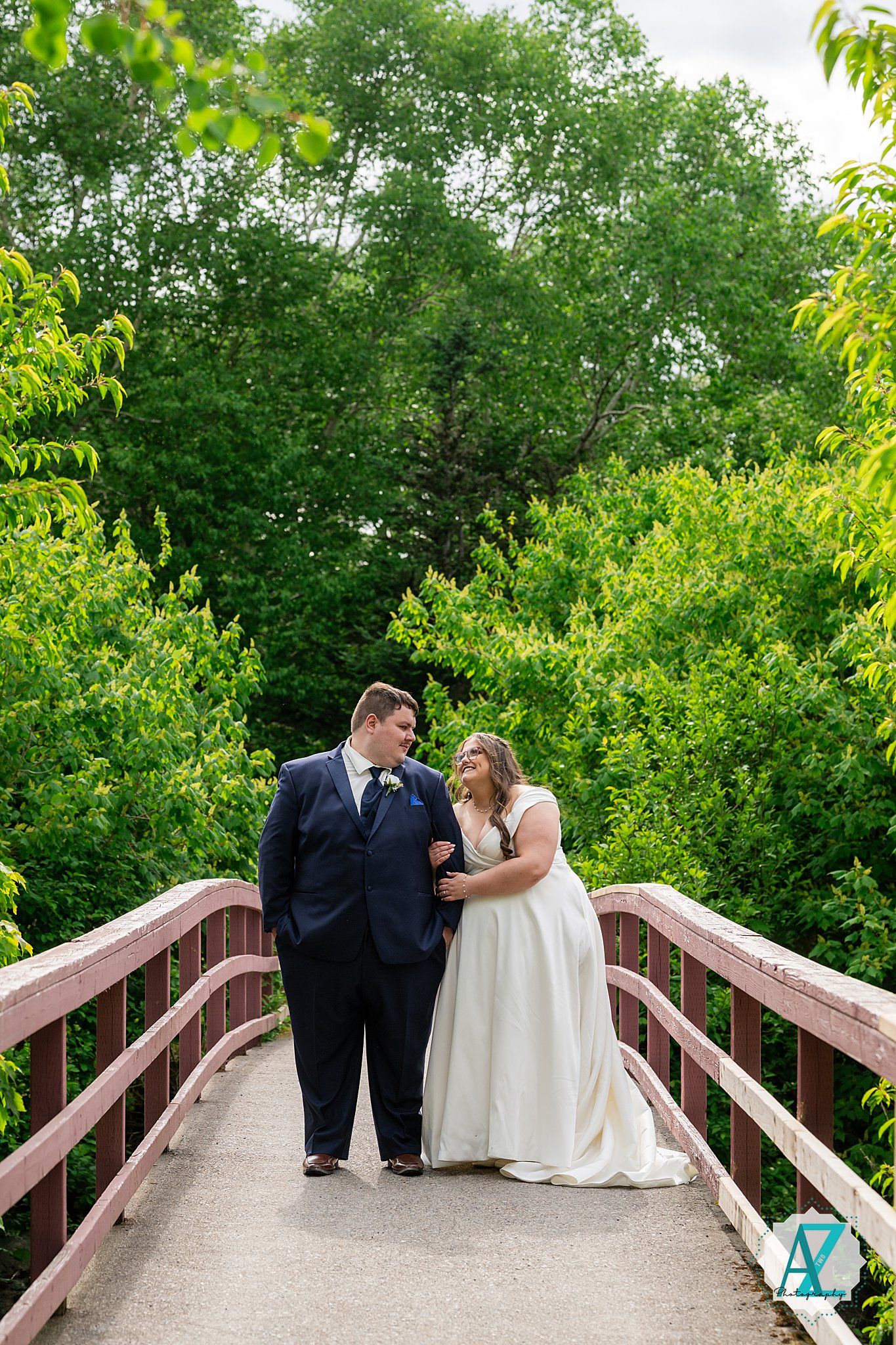 A couple in wedding attire shares intimate moments on a wooden footbridge surrounded by lush green foliage.