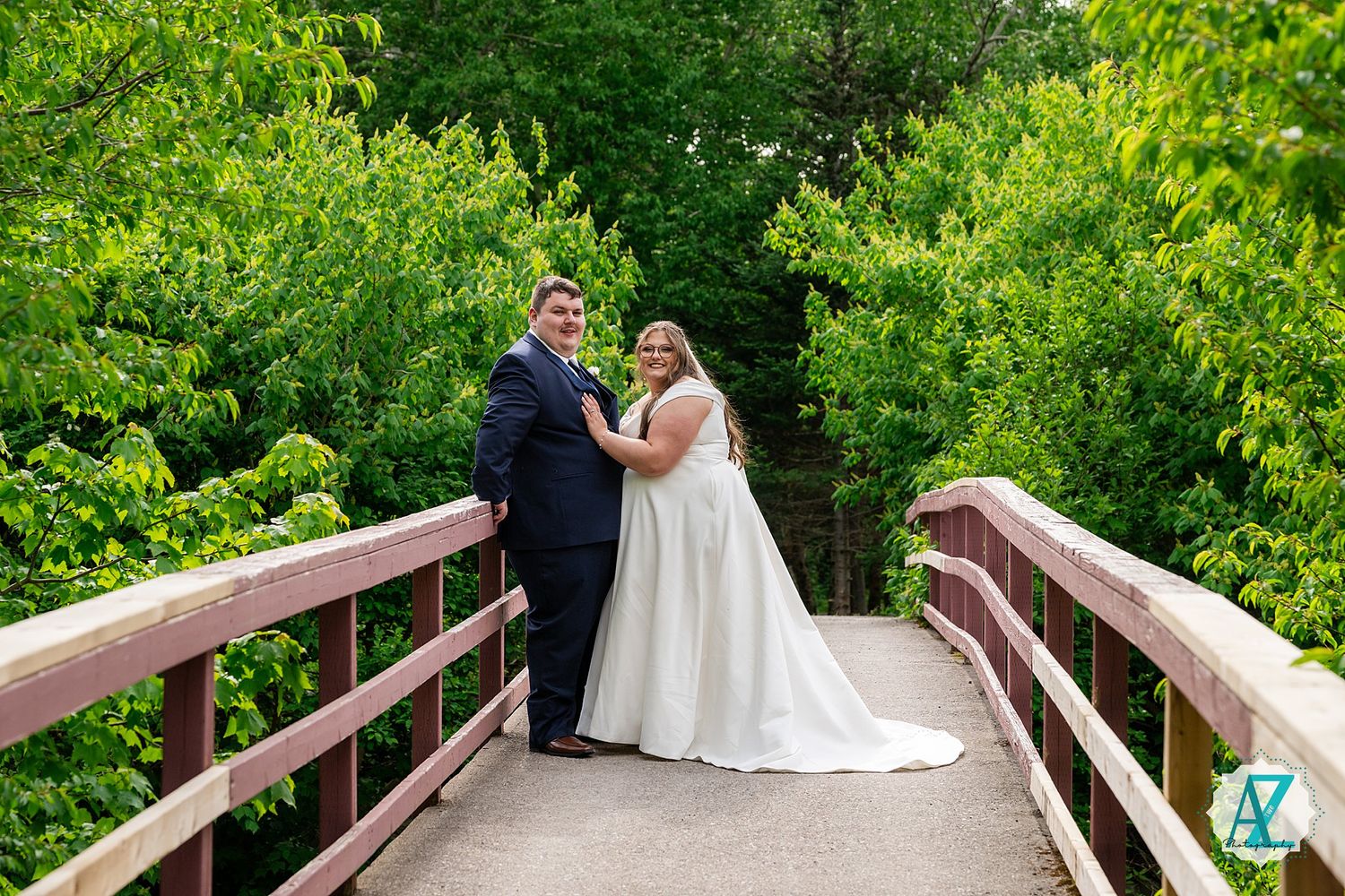 Couple embraces on a wooden footbridge surrounded by lush green summer foliage in romantic outdoor setting.