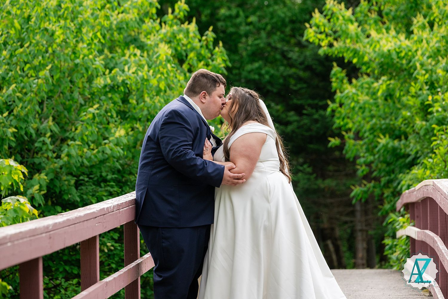 Two people share a tender moment while embracing on a wooden bridge surrounded by lush green foliage.