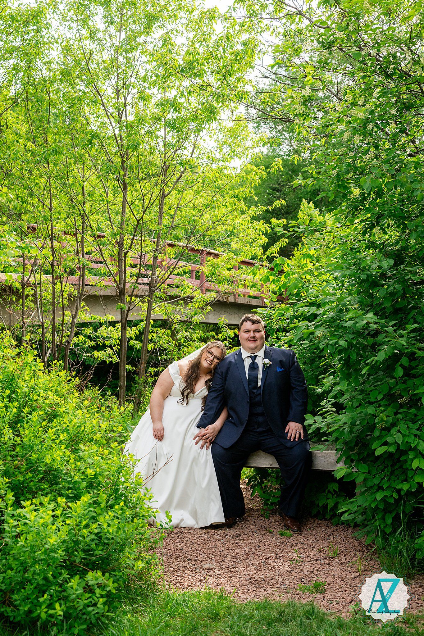 A newlywed couple shares a tender moment on a rustic wooden bridge surrounded by lush green foliage in a garden setting.
